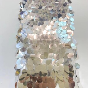 May include: A white fabric with silver sequins in a graduated pattern. The sequins are oval shaped and have a slight iridescent sheen.