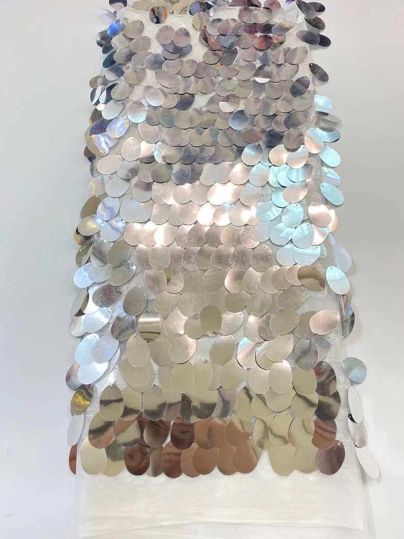 May include: A white fabric with silver sequins in a graduated pattern. The sequins are oval shaped and have a slight iridescent sheen.