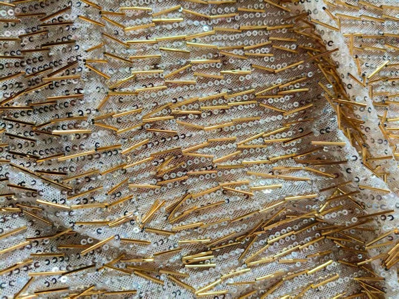 May include: A sheer fabric featuring a dense pattern of small, round, iridescent white sequins and elongated, metallic gold-toned bugle beads arranged in a scattered, multi-directional layout across the entire surface of the material.