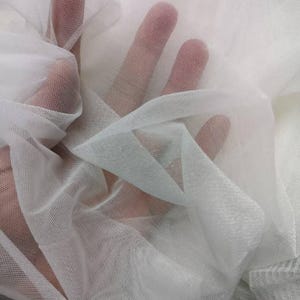 May include: Close-up of sheer white fabric, possibly tulle or netting, with a fine mesh texture. The fabric is draped and layered, creating shadows and highlights. The image shows the fabric's transparency and delicate nature.