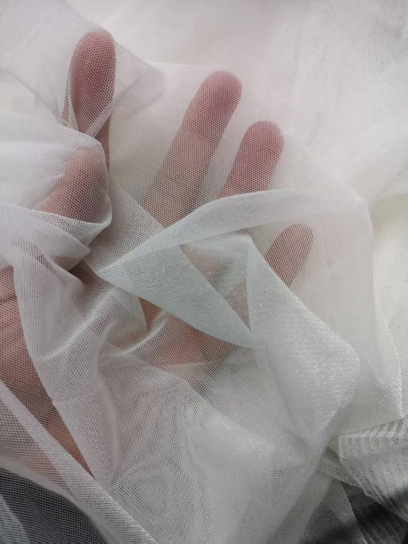 May include: Close-up of sheer white fabric, possibly tulle or netting, with a fine mesh texture. The fabric is draped and layered, creating shadows and highlights. The image shows the fabric's transparency and delicate nature.