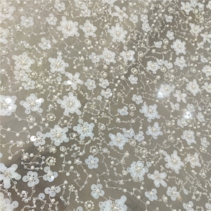 Könnte beinhalten: Weißer Blumenspitzenstoff mit Pailletten und Perlen. Der Stoff hat ein zartes, filigranes Design mit kleinen weißen Blüten und Perlen, die über den Stoff verteilt sind.