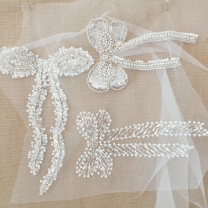 May include: Three white beaded bow embellishments on a sheer white fabric background. The bows are made of white beads and sequins.