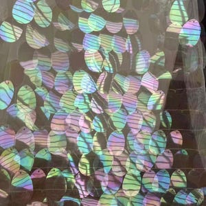 May include: Close-up of iridescent sequins in various shapes, including circles and teardrops, sewn onto a sheer fabric. The sequins reflect a rainbow of colors, creating a shimmering effect. The fabric is likely used for embellishment or decoration.