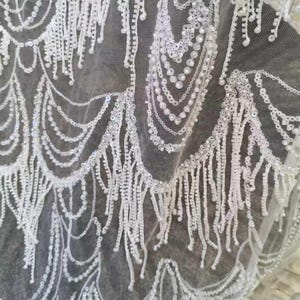 May include: Close-up of white lace fabric with intricate beaded and sequined patterns. The design features scalloped edges and dangling fringe, creating a delicate and textured appearance. Suitable for bridal wear or special occasion garments.