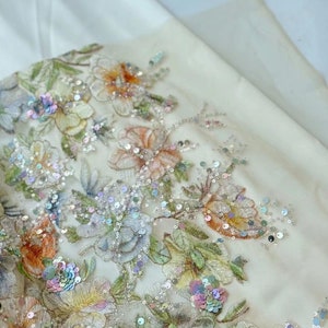 May include: Close-up of a white fabric with embroidered floral designs and iridescent sequins. The fabric features a variety of flowers in shades of pink, yellow, and orange.