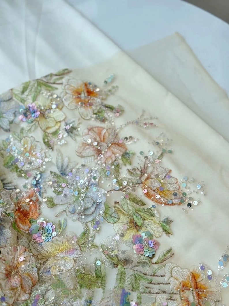 May include: Close-up of a white fabric with embroidered floral designs and iridescent sequins. The fabric features a variety of flowers in shades of pink, yellow, and orange.