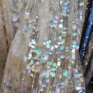 May include: A sheer fabric featuring rows of iridescent, circular sequins that reflect light with a rainbow-like sheen. The translucent mesh base is densely covered with these shimmering, reflective discs, creating a textured, light-catching surface.