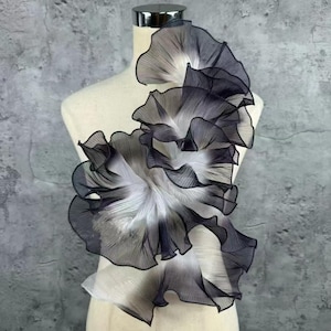 Puede incluir: Adorno de tela decorativo en capas en tonos negro, blanco y gris. El diseño presenta formas rizadas en forma de pétalos, creando una apariencia fluida y texturizada. La tela está adherida a un maniquí, lo que sugiere su uso en moda o diseño de vestuario.