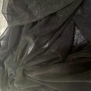 May include: Close-up of black, sheer fabric with a subtle shimmer. The fabric is layered and draped, creating soft folds and shadows. The texture appears delicate and lightweight, suitable for apparel or decorative purposes.