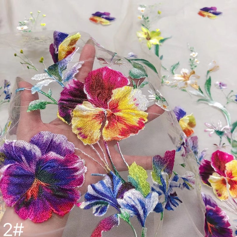 May include: Close-up of a sheer fabric with colourful embroidered flowers. The flowers are in shades of purple, yellow, red, and green. The fabric has a delicate, lacy texture.