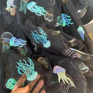 Pode incluir: Tecido de tule preto com desenhos de água-viva de lantejoulas iridescentes em tons de azul, verde, laranja e rosa. O tecido é drapeado, mostrando o trabalho detalhado das lantejoulas e a transparência do material.