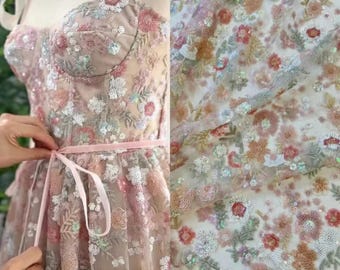 May include: A detailed view of a floral embroidered dress with a pink ribbon belt. The dress features a strapless bodice and a full skirt, adorned with white, pink, and peach-colored floral embroidery and sequins. The fabric is sheer and delicate.
