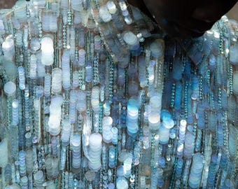 May include: A long-sleeved shirt covered in iridescent, light blue and white circular sequins and vertical strands of matching beads. The garment features a classic collar and a button-down front, with the sequins creating a reflective, shimmering effect.