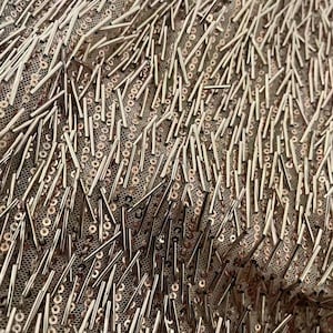 K&ouml;nnte beinhalten: Nahaufnahme eines Stoffes mit einer strukturierten Oberfl&auml;che. Der Stoff ist mit kleinen, metallischen Pailletten und l&auml;ngeren, metallischen Perlen bedeckt. Die Pailletten und Perlen sind in einem zuf&auml;lligen Muster angeordnet.