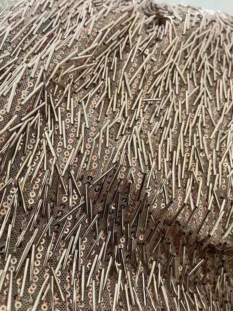 K&ouml;nnte beinhalten: Nahaufnahme eines Stoffes mit einer strukturierten Oberfl&auml;che. Der Stoff ist mit kleinen, metallischen Pailletten und l&auml;ngeren, metallischen Perlen bedeckt. Die Pailletten und Perlen sind in einem zuf&auml;lligen Muster angeordnet.