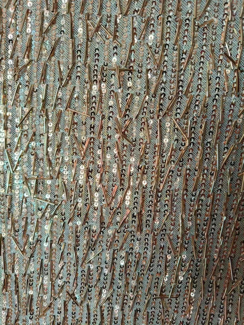 May include: A sheer fabric base featuring vertical rows of small, round, rose gold sequins interspersed with scattered, thin, metallic rose gold bugle beads arranged in a random, non-directional pattern across the entire surface of the material.