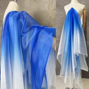 May include: A sheer fabric with a blue to white gradient. The fabric is draped over a mannequin, showcasing its flowing quality. The material appears lightweight and suitable for apparel or decorative purposes.