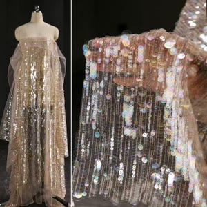Puede incluir: Un vestido sin tirantes hecho de tela de malla transparente cubierta de lentejuelas circulares iridiscentes que reflejan la luz en varios colores. El vestido presenta una silueta fluida con paneles laterales drapeados y una textura densa y brillante en toda la prenda.