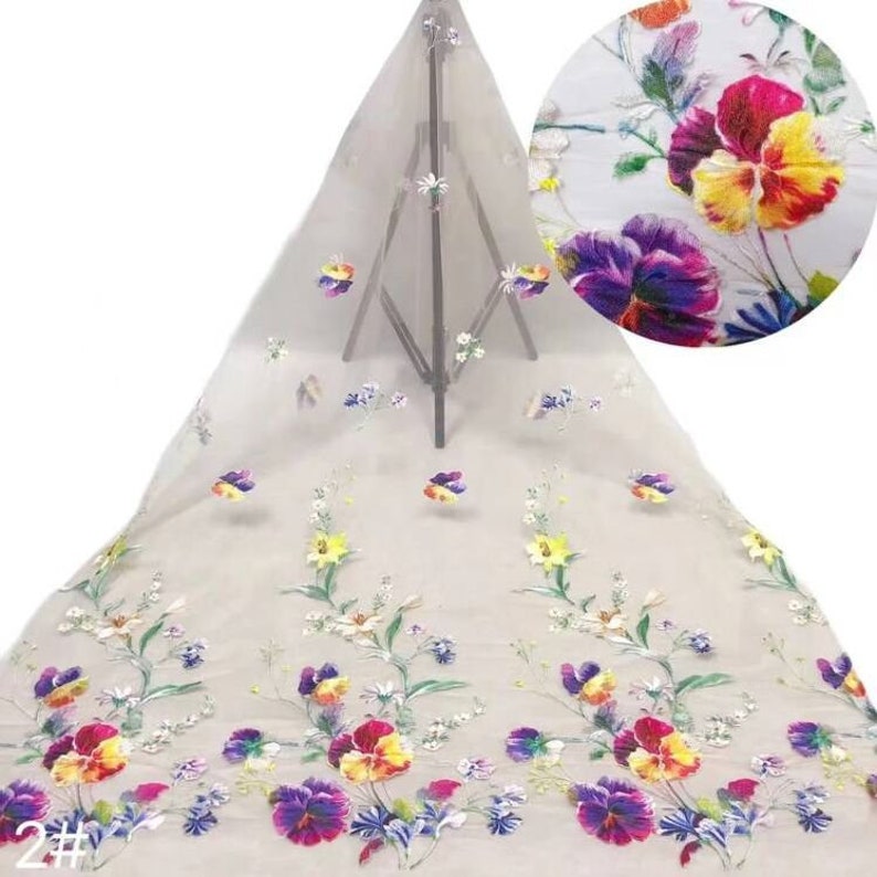 May include: White sheer fabric with colourful embroidered flowers. The flowers are a mix of pinks, yellows, blues, and purples. The fabric is delicate and has a soft texture.