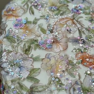 May include: Close-up of a floral embroidered fabric with iridescent sequins. The fabric features a delicate floral pattern with pastel colours and a sheer, mesh-like background.