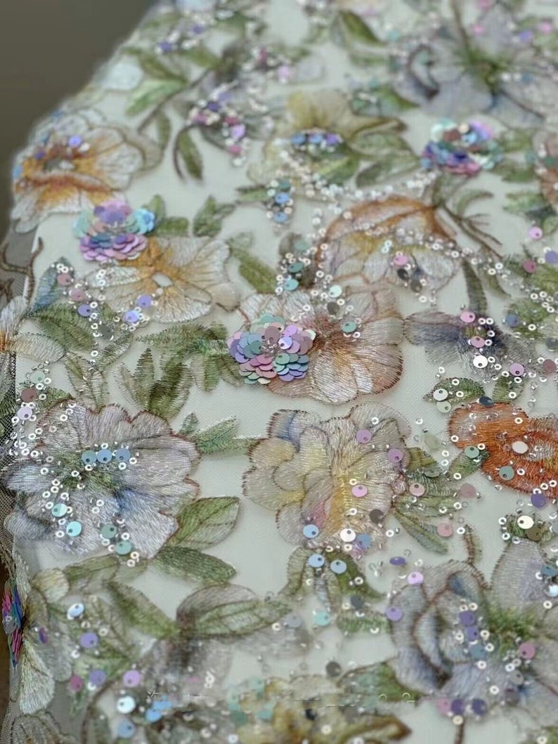 May include: Close-up of a floral embroidered fabric with iridescent sequins. The fabric features a delicate floral pattern with pastel colours and a sheer, mesh-like background.
