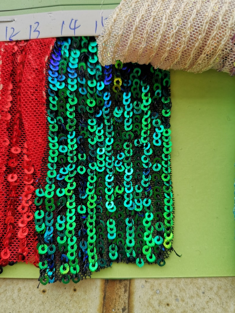 May include: A close-up of a green, blue, and black sequined fabric with a red sequined fabric to the left. The fabric is labelled with the numbers 12, 13, 14, and 15.