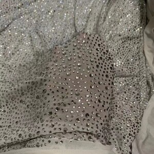 May include: Close-up of a silver fabric covered in numerous small, sparkling rhinestones. The fabric appears to be layered, with a sheer, white material visible underneath. The rhinestones catch the light, creating a shimmering effect.