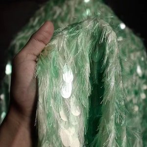 Puede incluir: Una tela de color verde menta que presenta una capa densa de flecos largos y finos junto con lentejuelas circulares reflectantes. El material tiene una apariencia texturizada y en capas con una calidad ligera, mostrando un color verde pálido.