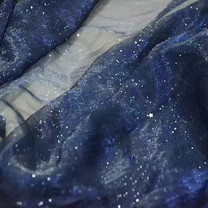 Può includere: Primo piano di un tessuto fluido con un colore di base blu scuro, quasi nero. Il tessuto è cosparso di piccoli punti bianchi scintillanti, simili a stelle. Il materiale ha una qualità traslucida, con aree blu e bianche più chiare.