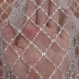 May include: Close-up of a sheer, white fabric with a diamond grid pattern. The grid is formed by rows of small, clear sequins. The fabric is slightly translucent, revealing the background. This material is suitable for bridal wear or decorative projects.
