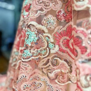 Puede incluir: Primer plano de una tela transparente con un diseño intrincado. La tela presenta una base rosa claro con patrones bordados en tonos de rosa, blanco y turquesa. El diseño incluye formas arremolinadas en forma de nube y está adornado con lentejuelas.