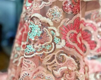 May include: Close-up of a sheer fabric with an intricate design. The fabric features a light pink base with embroidered patterns in shades of pink, white, and teal. The design includes swirling cloud-like shapes and is embellished with sequins.