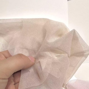 May include: Close-up of a sheer, light beige fabric with a fine mesh texture. The fabric is gathered and held, showing its delicate nature. The background is plain white, highlighting the fabric's color and texture.