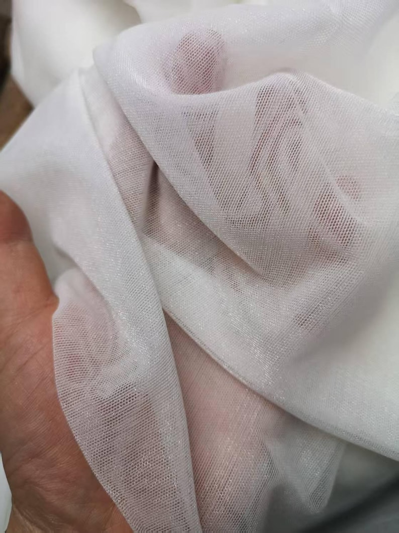 May include: Close-up of white sheer fabric. The fabric is thin and delicate, with a fine mesh weave. The light passes through the fabric, creating highlights and shadows. The fabric is soft and flowing, with a slight sheen.