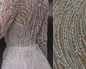 Puede incluir: Un vestido blanco con cuello alto y mangas largas, cubierto de adornos que parecen perlas. El vestido tiene un corpiño ajustado y una falda fluida.