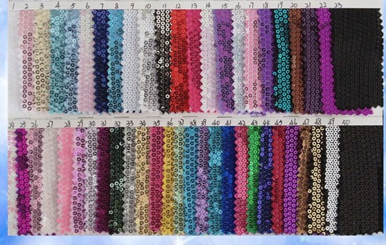Op de afbeelding: Een kleurstaal met 50 verschillende kleuren van paillettenstof. De stof is gerangschikt in rijen van 10, waarbij elke rij genummerd is van 1 tot 50. De kleuren vari&euml;ren van licht tot donker en omvatten tinten van goud, zilver, rood, blauw, groen, paars, roze en zwart.