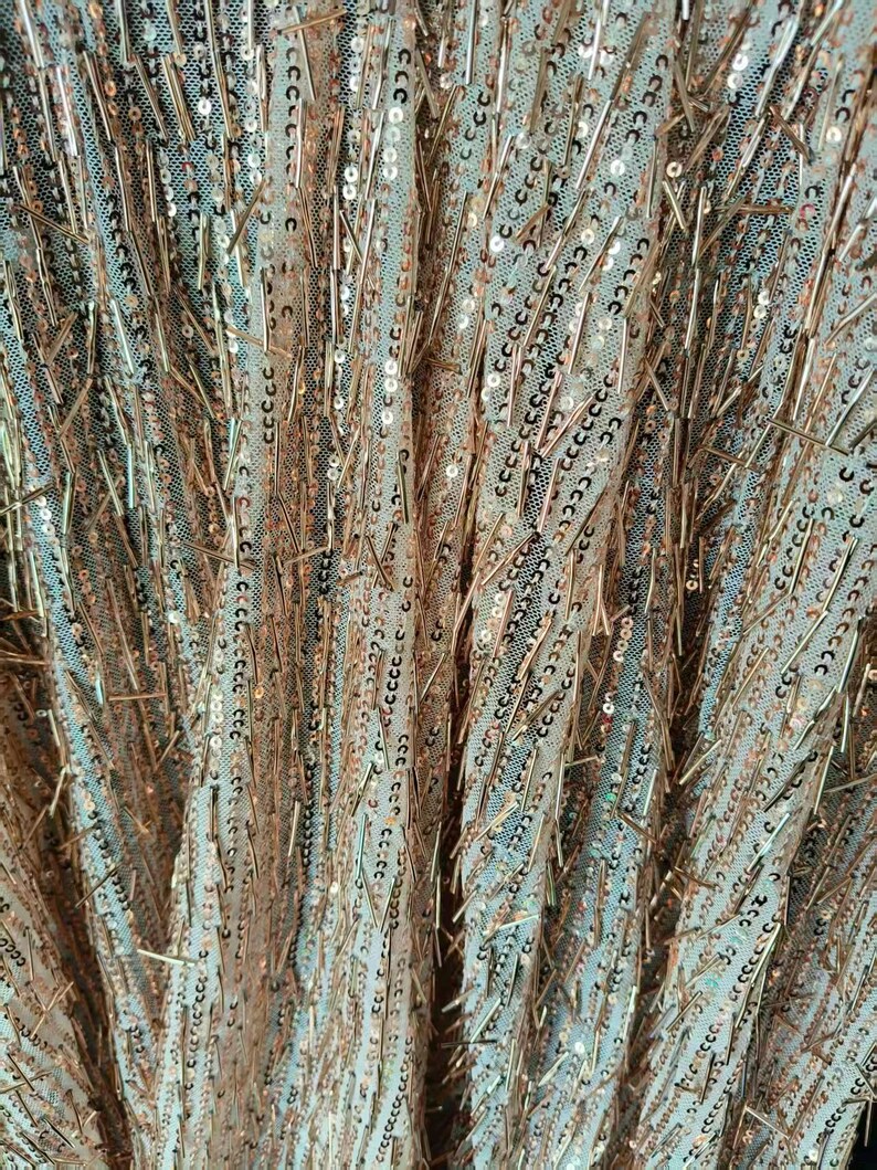 May include: A sheer fabric textile featuring a dense arrangement of rose gold sequins and thin, metallic rose gold bugle beads arranged in vertical, slightly irregular lines across a light-colored, fine mesh netting base material.