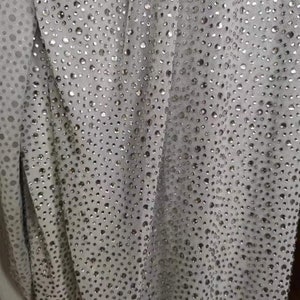 May include: Close-up of a light gray fabric covered in small, shiny, silver-colored embellishments. The fabric is draped, creating soft folds and shadows. The embellishments are evenly spaced across the material.