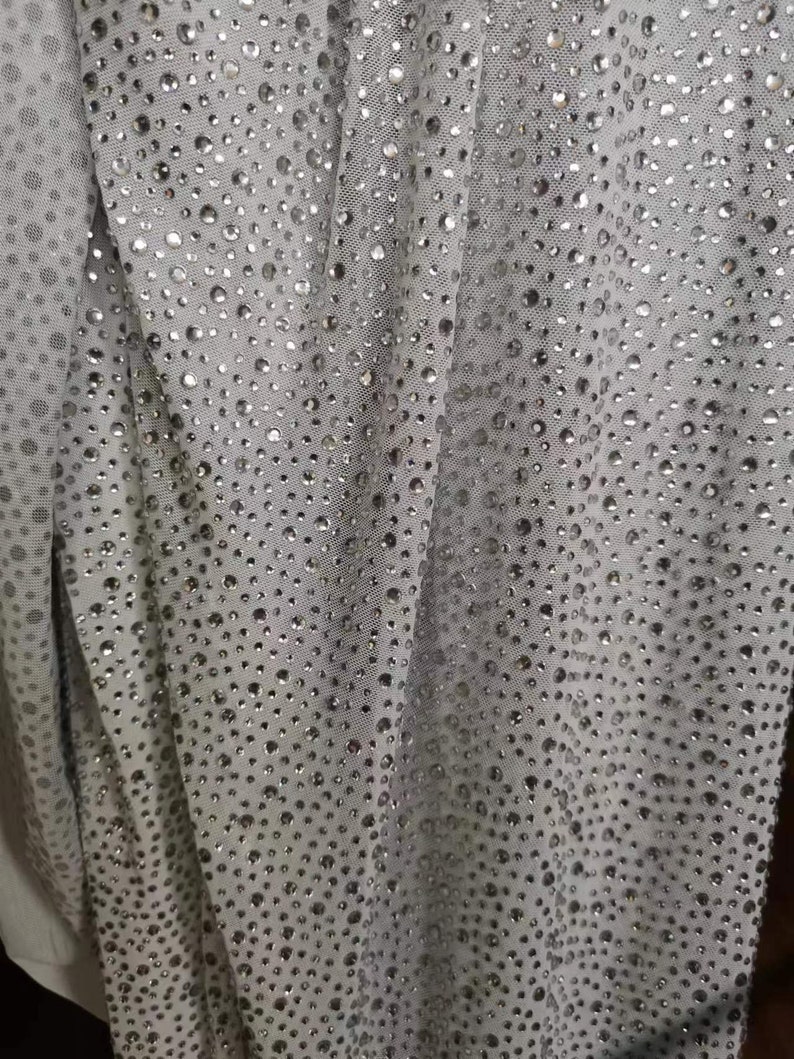 May include: Close-up of a light gray fabric covered in small, shiny, silver-colored embellishments. The fabric is draped, creating soft folds and shadows. The embellishments are evenly spaced across the material.