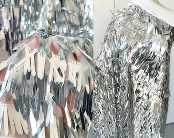 Könnte beinhalten: Silberner Fransenbesatzstoff mit metallischem Glanz. Die Fransen bestehen aus dünnen, flachen Stoffstreifen, die zusammengenäht sind, um einen fließenden, kaskadierenden Effekt zu erzeugen.