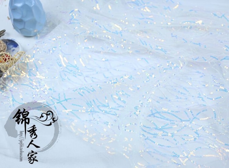 May include: Light blue iridescent sequins on a sheer mesh fabric. The sequins are arranged in a wavy pattern.