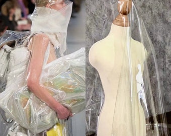 May include: A fashion-forward image featuring a person in a clear plastic garment and a dress form draped with clear plastic. The person is holding a large clear plastic bag. The dress form is beige with a wooden neck.