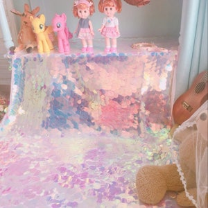 May include: A white iridescent sequin tablecloth with a pink, blue, and purple shimmer.  Three dolls with pink dresses and brown hair are sitting on the tablecloth.  A yellow pony and a brown deer are also on the tablecloth.
