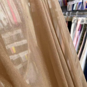 May include: Close-up of a shimmering, sheer fabric in a warm, golden-beige hue. The fabric drapes in soft folds, catching the light and creating a subtle sparkle. The background shows a glimpse of fabric samples, suggesting a textile shop.