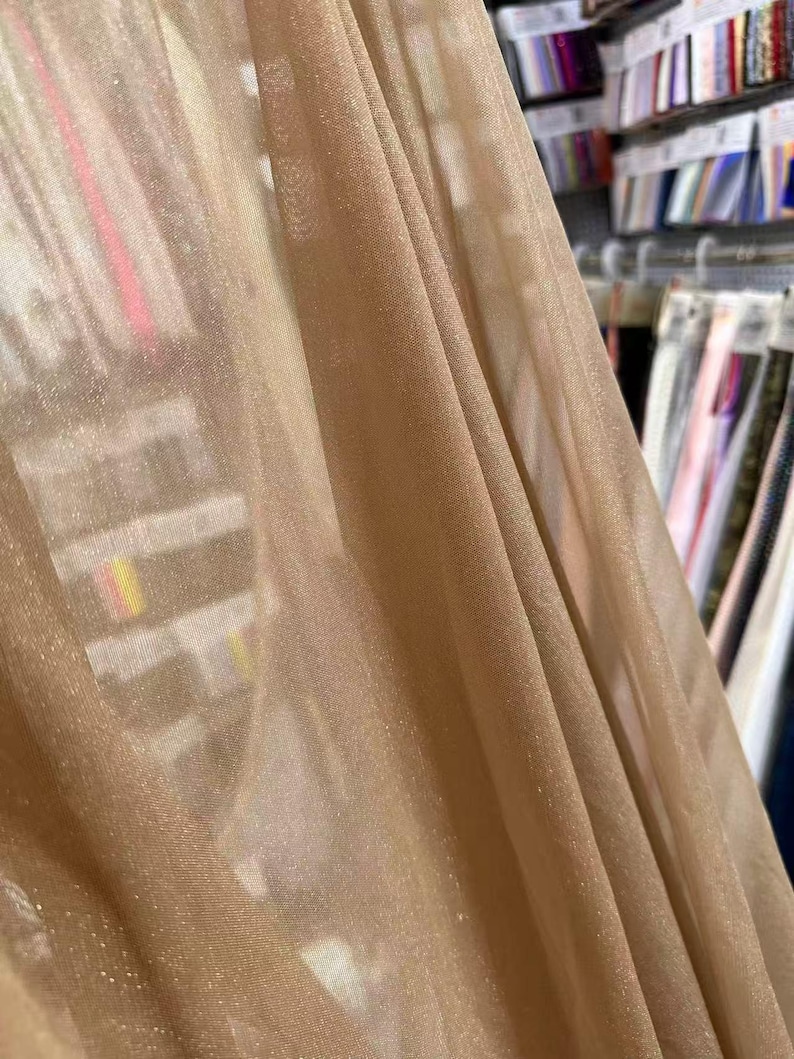 May include: Close-up of a shimmering, sheer fabric in a warm, golden-beige hue. The fabric drapes in soft folds, catching the light and creating a subtle sparkle. The background shows a glimpse of fabric samples, suggesting a textile shop.