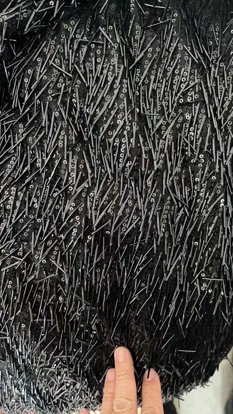 K&ouml;nnte beinhalten: Schwarzer Stoff mit einer strukturierten Oberfl&auml;che, bedeckt mit kleinen schwarzen Pailletten und langen schwarzen F&auml;den.