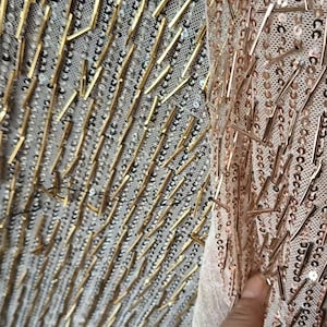 May include: A sheer fabric textile featuring a dense pattern of metallic gold-toned cylindrical beads and small circular sequins. The embellishments are arranged in vertical lines across the mesh base, creating a textured and reflective surface.