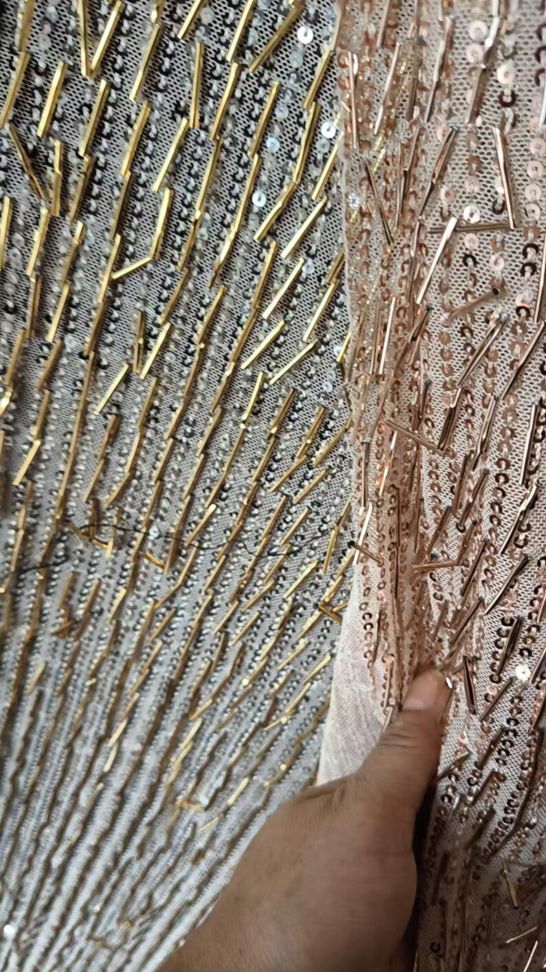 May include: A sheer fabric textile featuring a dense pattern of metallic gold-toned cylindrical beads and small circular sequins. The embellishments are arranged in vertical lines across the mesh base, creating a textured and reflective surface.