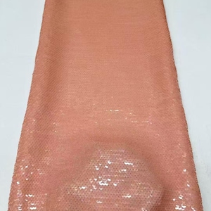 Puede incluir: Un trozo rectangular de tela cubierto de lentejuelas pequeñas y redondas de color melocotón. Las lentejuelas están densamente empaquetadas, creando un efecto brillante. La tela tiene una apariencia suave y fluida, adecuada para prendas o proyectos decorativos.
